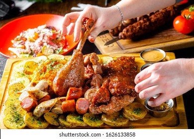 Background Black Burnt Carbonized Forest, Structure, Shot From The Side, Female Hand Corrects The Fried Leg Of A Turkey Thigh On A Cutting Board, Fried Potatoes And Tomatoes