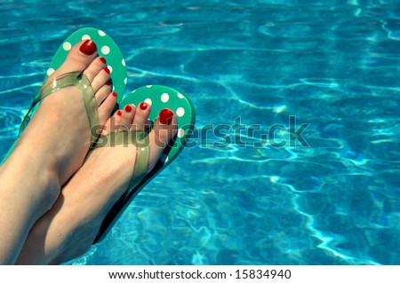 Background is filled with aqua water.  Woman has on polka dotted flip flops and has red painted toe nails.  Feet only.