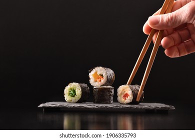 Background hands picking four assorted vegetable maki sushi with chopsticks on slate serving plate on dark wooden table with black background. Front view. Horizontal composition.