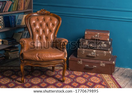 Background image of vintage old room with a bookshelf, leather and wooden armchair in classic style, colorful antique carpet and a lot of suitcases with blue wall. Copy space. Vogue and luxury.