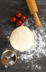 Background For Making Pizza, Raw Dough, Cherry Tomatoes And A Rolling Pin For Rolling Out The Dough On A Dark Background. Pizza Cooking Concept. Focus On The Dough