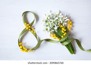 Background postcard for Women's day march 8, eight from a green ribbon and a bouquet of snowdrops, mimosa on a white wooden background