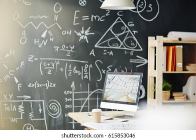 Background shot of empty workplace in sunlight: desk with computer and supplies standing against blackboard with scientific formulas written in chalk