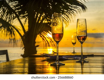 Background Of Three 3 Glasses Champagne Vine Staying On Wood Table Chairs Against Palm Silhouette Sunset Yellow Sky With Clouds Couple In Water On Coast And Sun Light Reflection On Boards After Dinner