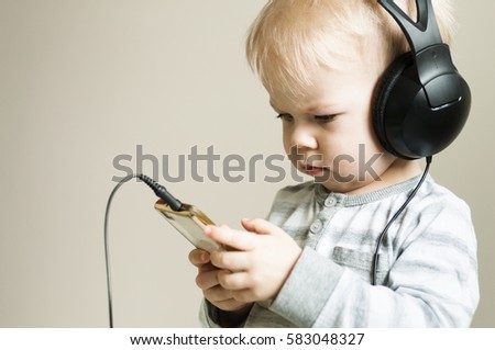 Background with two years old boy heeds in to the sounds of music in headphones, holding in hands the smartphone. Half profile.