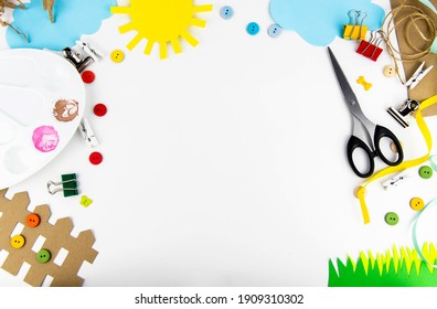 The background is a view from above on a white table with children's creativity. On the table of colored paper the sun, clouds, grass, fence, palette, scissors, colored buttons