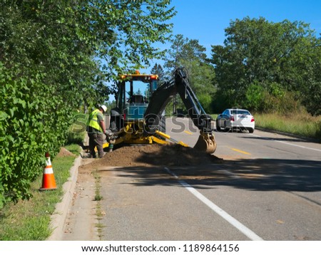 A backhoe and construction worker placing cables under street as car drives by