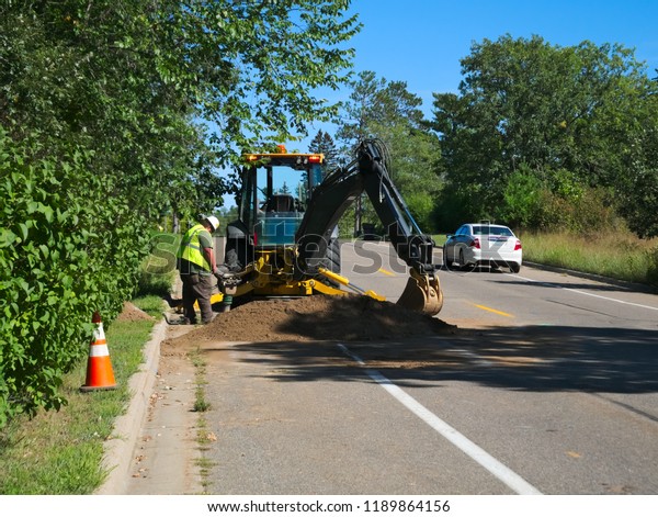 A backhoe and construction worker placing\
cables under street as car drives\
by
