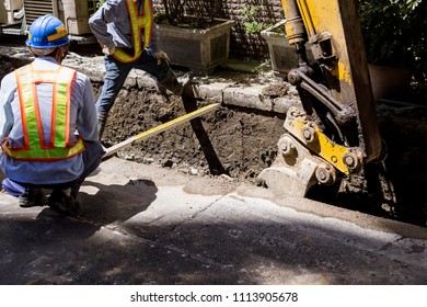 A Backhoe And Crew Dig A Utility Trench For Gas And Other Electrical Utilities At A Local Community. Workers Are Measuring The Width Of The Trench.