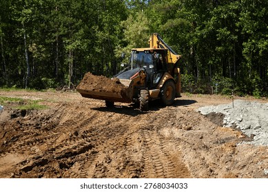 Backhoe loader carrying and dumping soil at a construction site, shaping ground and preparing foundation area in a forested residential location. 
