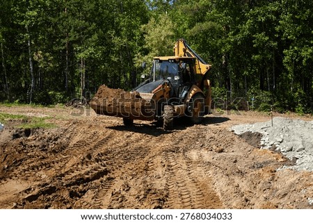 Backhoe loader carrying and dumping soil at a construction site, shaping ground and preparing foundation area in a forested residential location. 