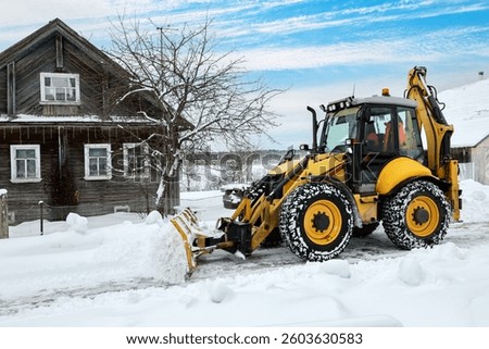 Backhoe loader with hydraulic front-mounted snow blade clears compacted snow using precision controls while reinforced tires ensure grip and stability on uneven icy rural terrain near buildings.