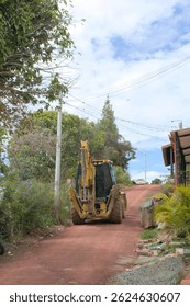 carregador retroescavadeira está posicionado em uma estrada de terra. A parte traseira do veículo está voltada para a câmera. Árvores e folhagem cercam o veículo, e linhas de energia são visíveis por cima.