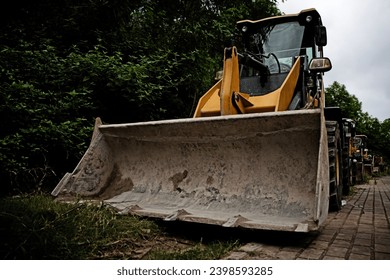 Backhoe loader, trees, paving stones, Tractor parked near the forest. Yellow tractor, excavator - heavy-duty machine.