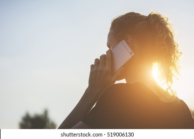 Backlight, Rear View, On Right Side Of Screen Young Woman, Dressed In Black T-shirt Talking On Cell Phone. In Background Blue Sky.Girl Using Mobile Phone, Digital Gadget