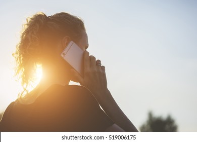 Backlight, Rear View, Young Woman, Dressed In A Black T-shirt Talking On A Cell Phone. In Background Blue Sky.Girl Using Gadget.Sunset. Summer Evening.