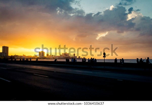 backlight of a\
street, on the edge of the sea, full of people with the sunset and\
the historic city in the\
background