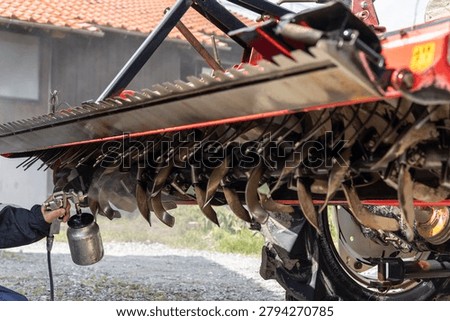 Backlit maintenance work spraying rust preventive oil onto the rotary tiller blades of a tractor in a rural spring setting