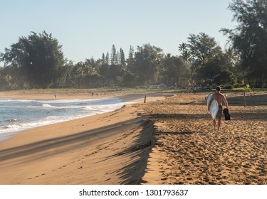 Backlit man carrying surf board across the sandy beach to the ocean in Kauai Hawaii