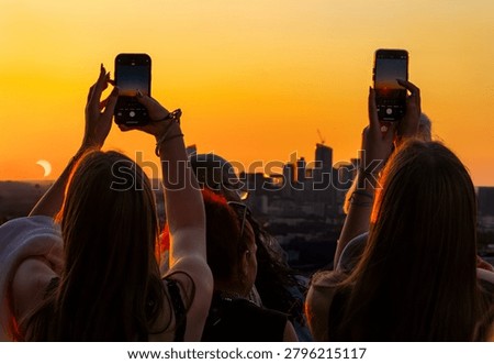 Backlit people, from the top of the Eiffel Tower, capturing a beautiful partial solar eclipse with their smartphones against a golden sunset sky with the Paris skyline and La Défense in the background