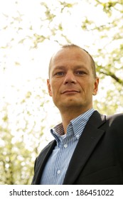 Backlit Portrait Of A Friendly Business Man. A Happy Average Guy, Dressed Smart Casual, Seen From A Low Point Of View, Looking Ito The Camera