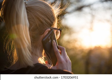 Backlit Rear View Of Young Woman Talking On Cell Phone Outdoors In Park At Sunset. Girl Holding Mobile Phone, Using Digital Device, Looking At Setting Sun. Copyspace.