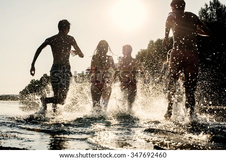 Backlit shot of teenagers running with their feet in the water at the beach during a sunny afternoon during summer break. They are having fun, playing and splashing water around them