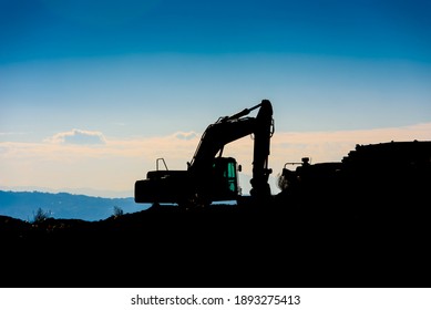Backlit silhouette of an excavator on top of a hill with unfocused sky background.