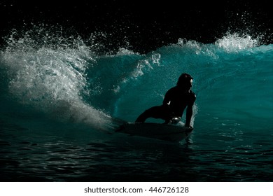 A backlit and silhouetted image of a surfer riding a blue wave at night.