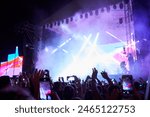 Backlit stage with vibrant lights at outdoor music festival. Fans record live performance on smartphones. Excitement in air, band plays hit songs. Summer night event, audience enjoys concert vibe.