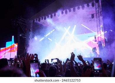 Backlit stage with vibrant lights at outdoor music festival. Fans record live performance on smartphones. Excitement in air, band plays hit songs. Summer night event, audience enjoys concert vibe.