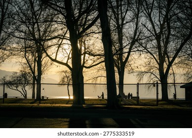 Backlit sunset through leafless trees at a lakeside park with fence and silhouettes of people by the water and distant mountains