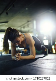 Backlit Vertical Shot Of Exhausted Young Woman Doing Forearm Plank Exercise During Workout In Gym