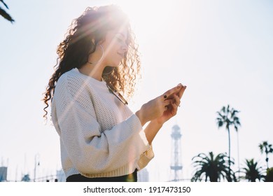 Backlit view of millennial blogger using cellphone technology for creating travel publication and share to social media during network browsing in city, curly hipster girl reading received message
