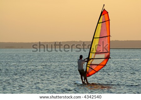 Backlit windsurfer at sunset on calm coastal water	
