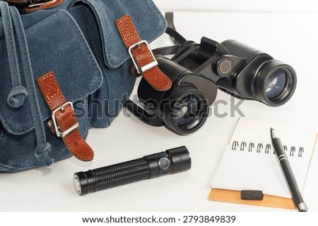 Backpack, binoculars, flashlight, and notebook in close-up against a white background.