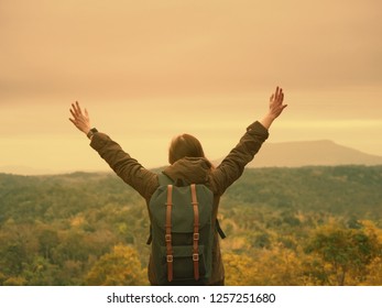backpack and camping concept from backside of long hair woman in warm cloth with her bag travel, relax, happy  and show her hand and see to forest and mountain in morning after sunrise