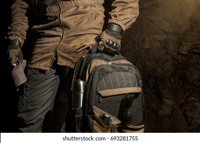 Backpack and gear composition.Man in storm jacket and tactical military gloves holding a backpack with travel gear on stone wall background.