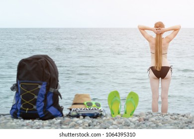 Backpack, hat and flip flops on the pebble beach. Girl in bikini and sea in the distance