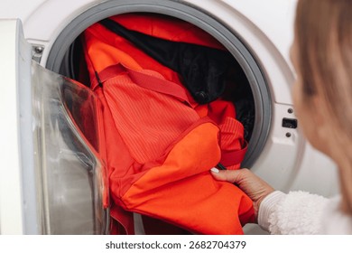 Backpack inside washing machine drum. Orange backpack during cleaning cycle. Household laundry concept with appliance. Domestic routine of washing bag. Hygiene care and cleaning process at home.
