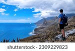 Backpack man with scenic view of Atlantic Ocean coastline and Anaga mountain range on Tenerife, Canary Islands, Spain, Europe. Looking at Roque de las Animas crag. Hiking trail from Afur to Taganana