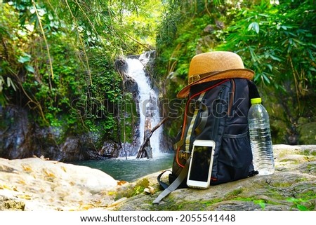 Backpack, a mobile phone and a bottle of water are laying on the rock by waterfall background for adventure and for hike in the forest
