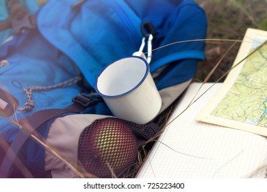 A backpack, a mug, a notepad and a map are lying on the grass. Tourist equipment. The apple lies in the pocket of the backpack. Walk in the forest. Camping in the mountains.