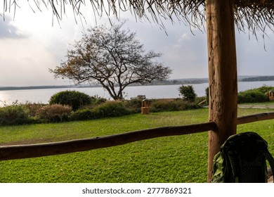 A backpack rests on the ground next to a wooden post while a tree stands by the lake in the background. The sun is setting, casting light over the water and grass.