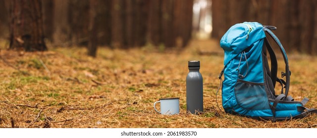 Backpack, thermos and travel mug in forest. Hiking equipment on footpath in woodland. Panoramic view