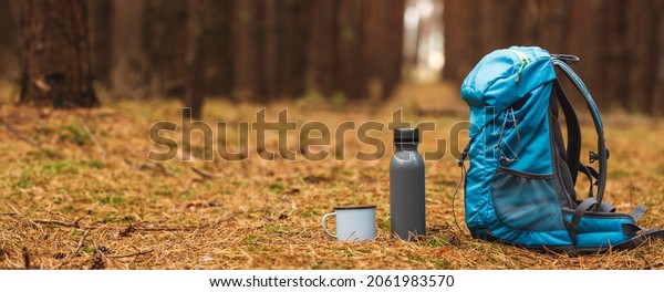 Backpack, thermos and travel mug\
in forest. Hiking equipment on footpath in woodland. Panoramic\
view