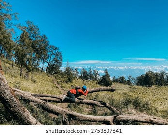A backpacker climbing over a fallen tree along the hiking trail of Mount Lawu via Candi Cetho in Karanganyar, Central Java, Indonesia. The scene shows a rugged mountain landscape with grassy hills, scattered highland trees, and a clear blue sky stretching across the horizon. In the distance, layers of white clouds create a dramatic backdrop, emphasizing the elevation and natural beauty of this volcanic region.

The traveler carefully crosses the large log that lies across the path, representing the small challenges often encountered during a trekking journey. Natural obstacles such as fallen trunks, rocky paths, and uneven terrain are common along this route, making every step part of an adventurous outdoor experience. The surrounding valley opens wide, revealing peaceful grassland and sparse forest typical of the upper slopes of Mount Lawu.

Bright daylight illuminates the environment, highlighting the textures of dry grass, wood, and soil along the trail. The wide landscape and open sky give a sense of freedom, exploration, and connection with nature. This moment captures the essence of mountain travel, where persistence and curiosity guide every movement toward the summit.

Located in Karanganyar, Central Java, the Candi Cetho route is known for its scenic views and unique highland ecosystem. The photograph portrays a quiet yet dynamic moment during a trek, reflecting adventure, endurance, and the rewarding feeling of exploring Indonesia’s mountainous wilderness.