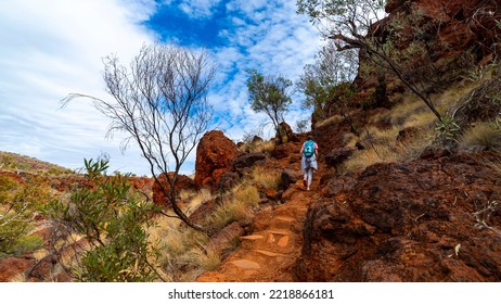 Backpacker Girl Hiking On Red Rocks Up A Steep Mountain In Karijini National Park In Western Australia, Desert Landscape Of Australian Outback