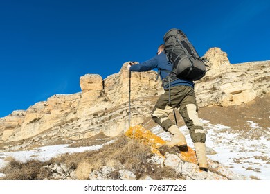 Backpacker with a large backpack and sticks ascends to the rock against the background of epic rocks in the winter season.
