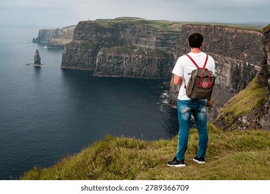 Backpacker standing on the edge of the Cliffs of Moher looking at the ocean scenery, Ireland
